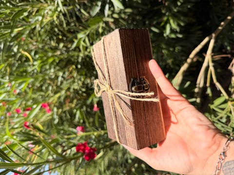 A hand holding the closed wooden keepsake box, tied with a jute twine bow, in front of green foliage.